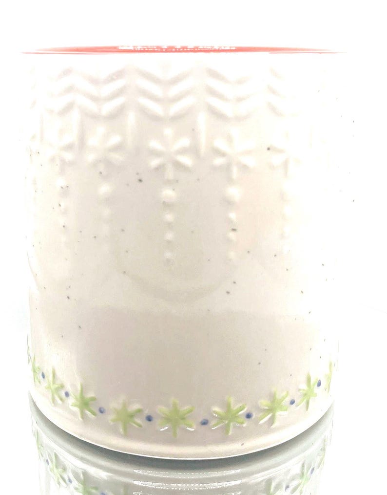 May include: A white ceramic mug with embossed snowflake and chevron patterns. The mug features a green and blue floral design around the base and a red rim. The mug is designed for beverages.
