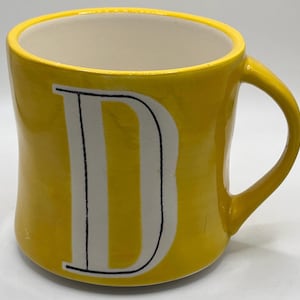 Puede incluir: Una taza de cer&aacute;mica amarilla con interior blanco y una gran letra "D" blanca en la parte delantera. La taza tiene un asa curva y un acabado brillante. La taza es de color amarillo brillante.