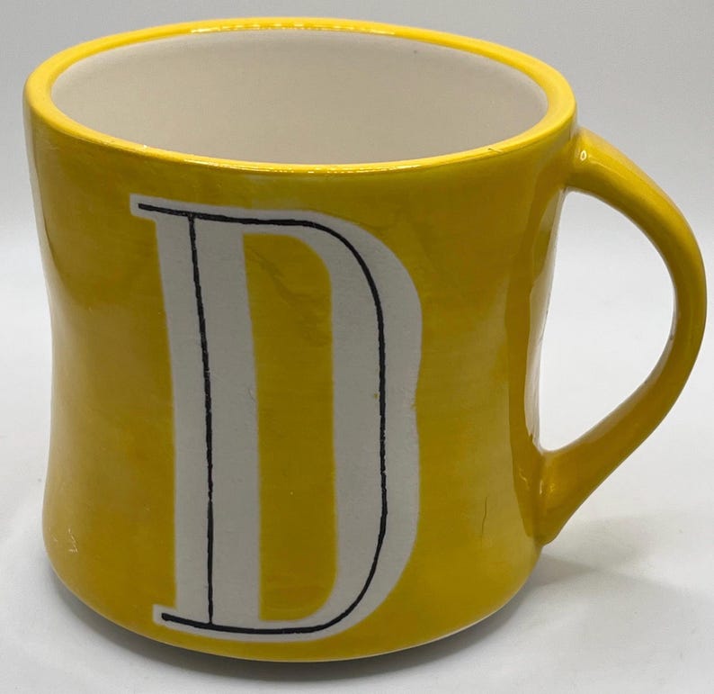 Puede incluir: Una taza de cer&aacute;mica amarilla con interior blanco y una gran letra "D" blanca en la parte delantera. La taza tiene un asa curva y un acabado brillante. La taza es de color amarillo brillante.
