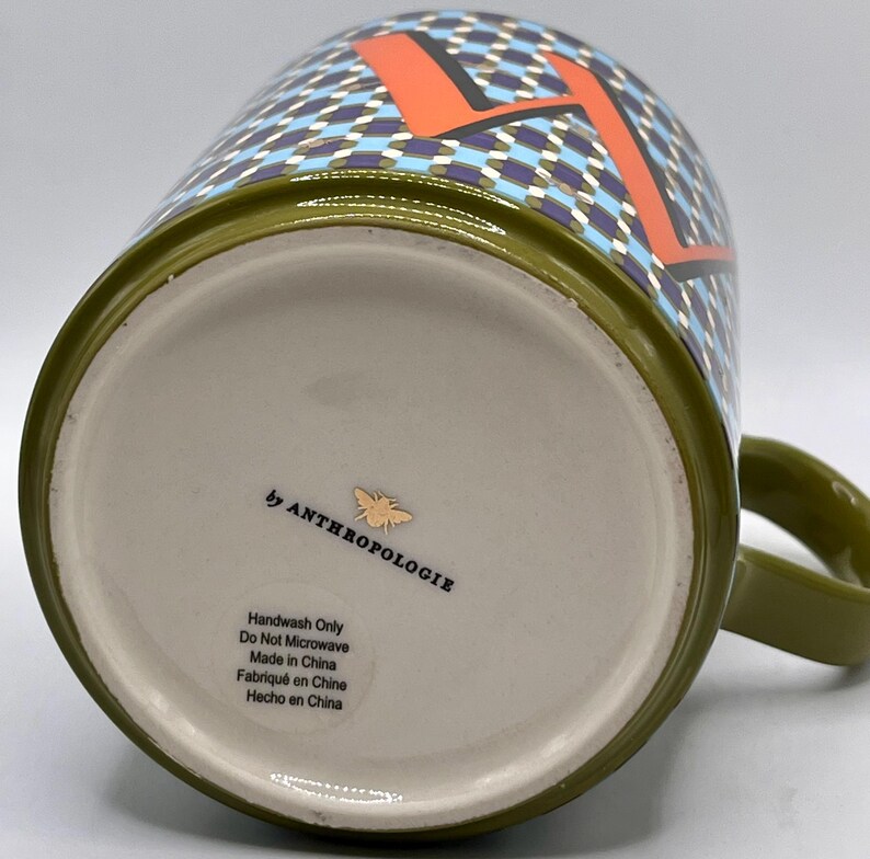 May include: Olive green ceramic mug with a handle. The mug features a geometric pattern in blue, purple, and white, with an orange letter "A". The bottom of the mug has the text "by ANTHROPOLOGIE" and care instructions.