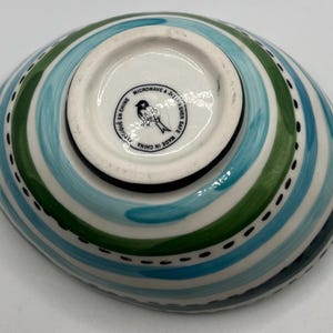 May include: An oval-shaped ceramic bowl with a colorful design. The bowl features alternating stripes of blue, green, and white, with a dotted black line along the edge. The bottom of the bowl has a white center with a bird logo and the text "Microwave & Dishwasher Safe Made in China".