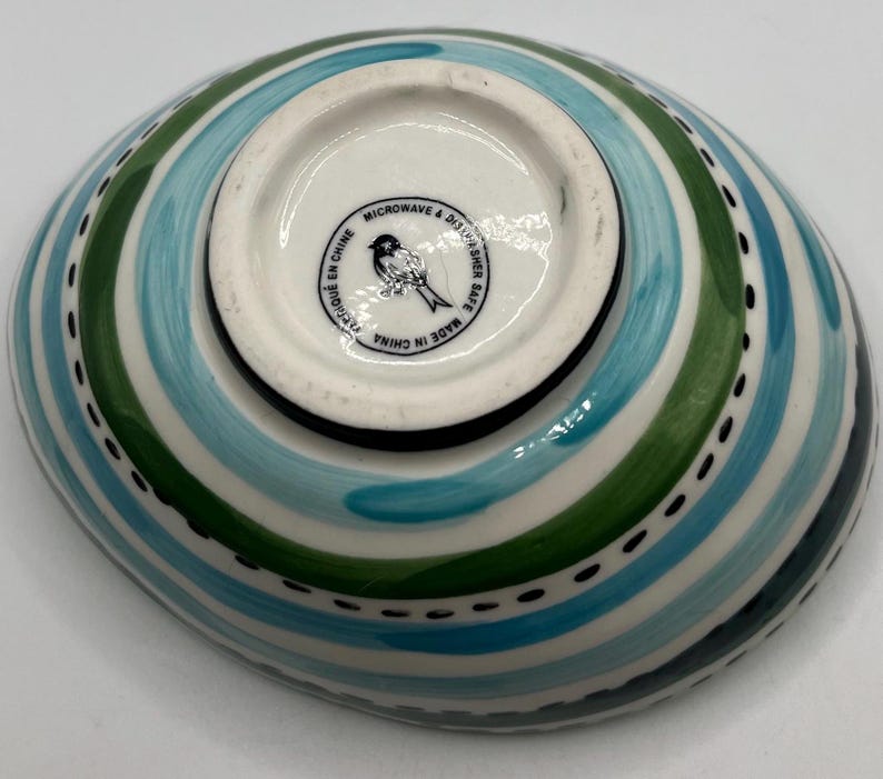 May include: An oval-shaped ceramic bowl with a colorful design. The bowl features alternating stripes of blue, green, and white, with a dotted black line along the edge. The bottom of the bowl has a white center with a bird logo and the text "Microwave & Dishwasher Safe Made in China".