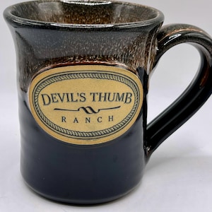May include: A ceramic mug with a dark blue base and a textured brown top. The mug features a label with the text "Devil's Thumb Ranch" in a decorative oval. The handle is black.