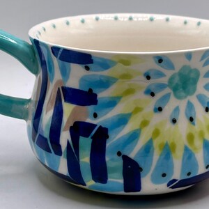 May include: A ceramic mug with a turquoise handle and a white interior. The exterior is decorated with blue, green, and brown abstract patterns. Small black dots encircle the rim.