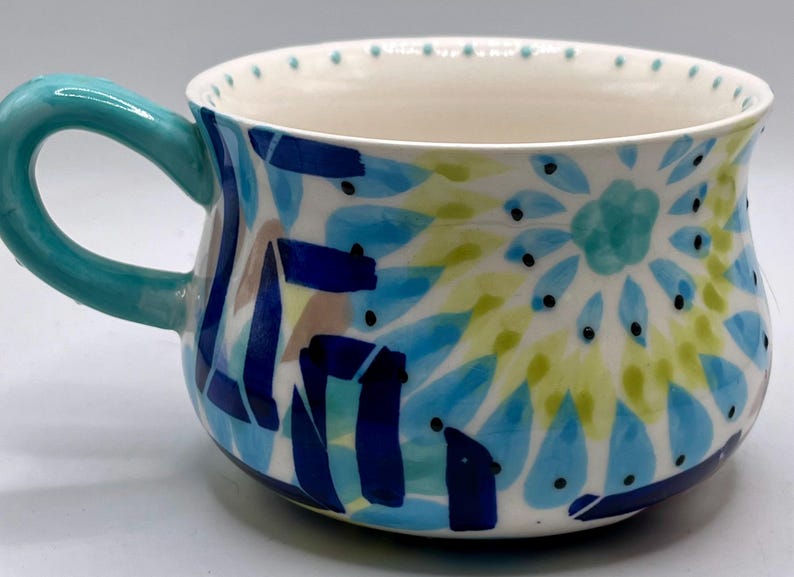 May include: A ceramic mug with a turquoise handle and a white interior. The exterior is decorated with blue, green, and brown abstract patterns. Small black dots encircle the rim.