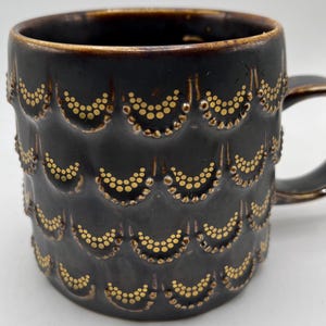 May include: A black ceramic mug with a gold pattern of small circles arranged in a scalloped design. The mug has a handle.