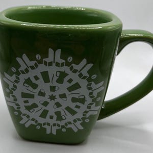 Op de afbeelding: Een groene keramische koffiemok met een wit ontwerp van koffiekopjes en koffiekannen die in een cirkel zijn gerangschikt. De mok heeft een handvat.