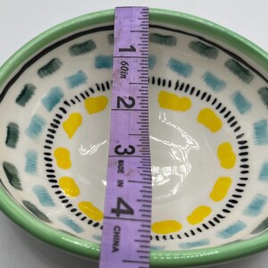 May include: A hand-painted ceramic bowl with a green rim, white interior, and colorful geometric patterns. The bowl's interior features blue, yellow, and black accents. The bowl is approximately 4 inches in diameter.