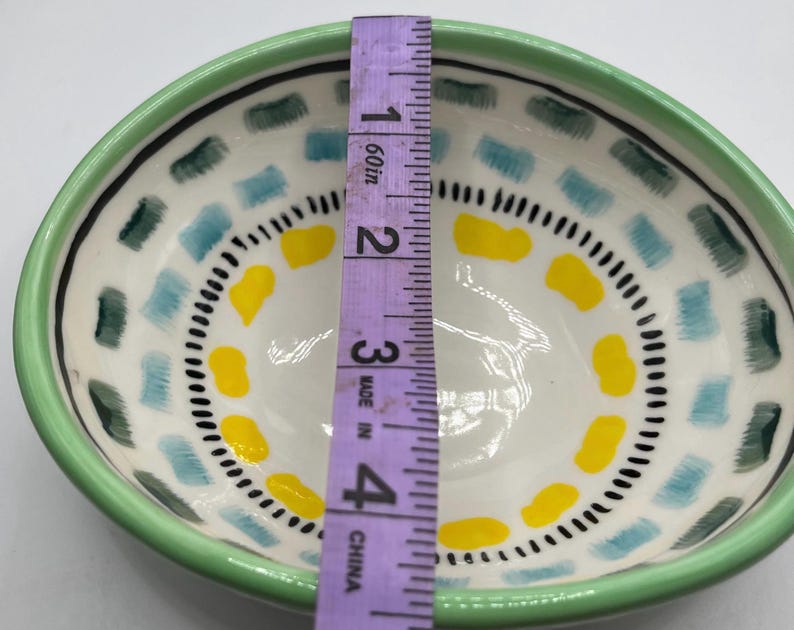 May include: A hand-painted ceramic bowl with a green rim, white interior, and colorful geometric patterns. The bowl's interior features blue, yellow, and black accents. The bowl is approximately 4 inches in diameter.