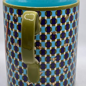 May include: A ceramic mug with a geometric pattern in blue, navy, and white, adorned with gold-colored bee illustrations. The mug features a teal interior and a green handle and rim.