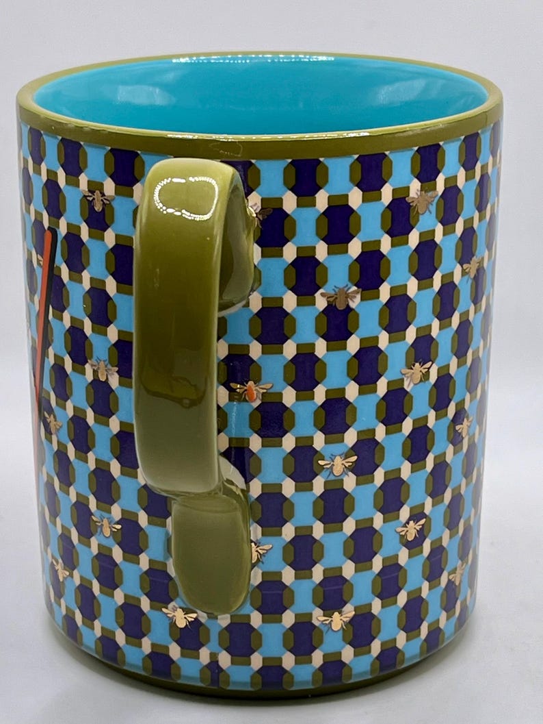 May include: A ceramic mug with a geometric pattern in blue, navy, and white, adorned with gold-colored bee illustrations. The mug features a teal interior and a green handle and rim.