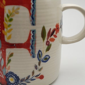 May include: White ceramic mug with a red letter "F" and floral designs in shades of blue, green, yellow, and orange.