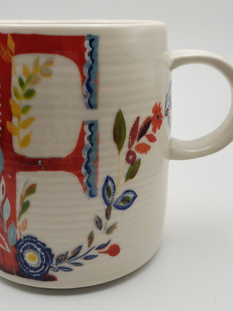 May include: White ceramic mug with a red letter "F" and floral designs in shades of blue, green, yellow, and orange.