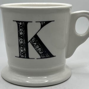 Può includere: Tazza in ceramica bianca con una lettera K nera sul davanti. La lettera K ha un design decorativo.