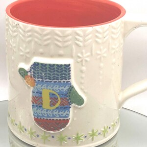 May include: Cream-colored ceramic mug with a red interior and a white handle. The mug features a raised mitten design with a yellow "D" and colorful patterns. The exterior has embossed leaf and snowflake patterns, with green star accents at the base.