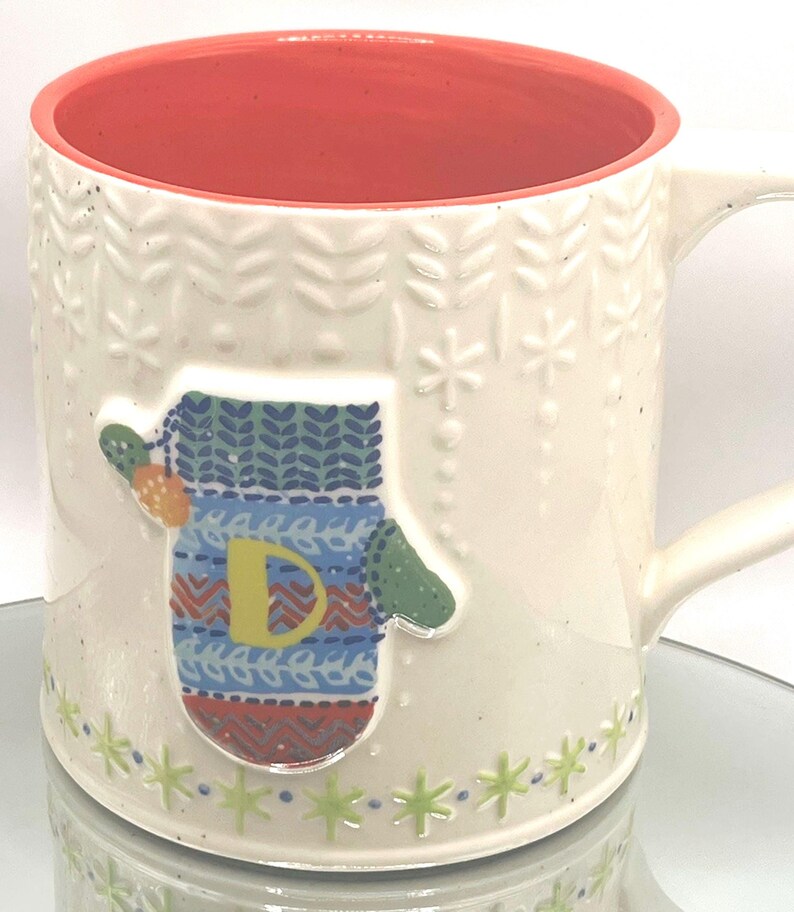 May include: Cream-colored ceramic mug with a red interior and a white handle. The mug features a raised mitten design with a yellow "D" and colorful patterns. The exterior has embossed leaf and snowflake patterns, with green star accents at the base.