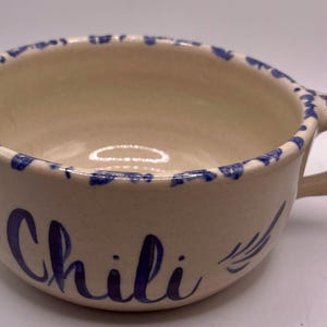 Może przedstawiać: Kremowa miska ceramiczna z jednym uchwytem, z napisem Chili namalowanym niebieską czcionką z przodu. Krawędź i uchwyt ozdobione są niebieskim wzorem malowanym gąbką, a wnętrze ma gładkie, błyszczące wykończenie o jasnym odcieniu.