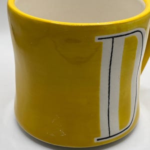 Puede incluir: Una taza de cer&aacute;mica amarilla con interior blanco y asa curva. El exterior presenta una gran letra "D" blanca con contornos negros y rayas verticales. La taza tiene un acabado brillante.