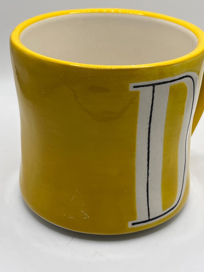 Puede incluir: Una taza de cer&aacute;mica amarilla con interior blanco y asa curva. El exterior presenta una gran letra "D" blanca con contornos negros y rayas verticales. La taza tiene un acabado brillante.