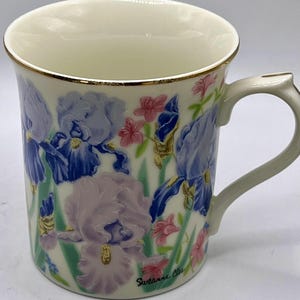 May include: Cream-colored ceramic mug with a gold rim and handle. The mug is decorated with embossed blue, purple, and pink iris flowers and green leaves. The artist's signature, "Suzanne Clee," is visible on the mug.