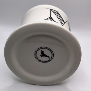 Puede incluir: Taza de cer&aacute;mica blanca con un dise&ntilde;o de la letra "M" en negro. La taza tiene una base ancha y un cuerpo cil&iacute;ndrico. La parte inferior de la taza presenta un logotipo de p&aacute;jaro negro y el texto "Dishwasher & Microwave Safe Made in China Fabriqu&eacute; en Chine."