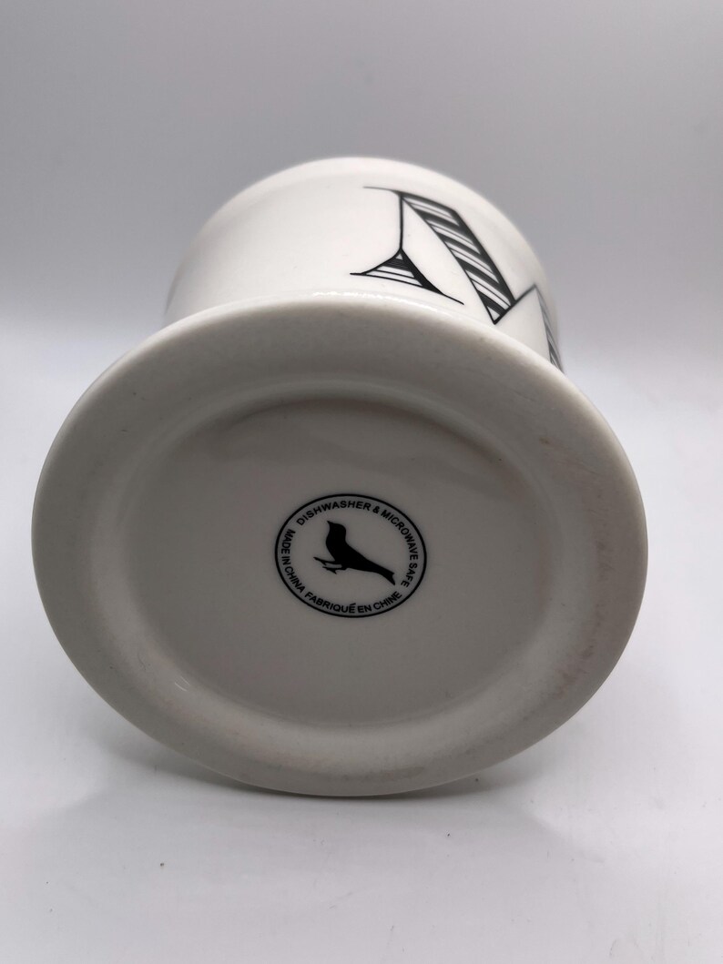 Puede incluir: Taza de cer&aacute;mica blanca con un dise&ntilde;o de la letra "M" en negro. La taza tiene una base ancha y un cuerpo cil&iacute;ndrico. La parte inferior de la taza presenta un logotipo de p&aacute;jaro negro y el texto "Dishwasher & Microwave Safe Made in China Fabriqu&eacute; en Chine."