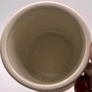 May include: Cream-colored ceramic mug with a rounded interior and a white exterior. The mug has a simple, classic design, suitable for hot beverages. The handle is visible on the right side of the mug.