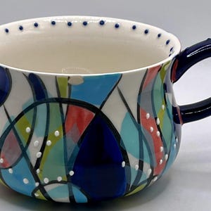 Może przedstawiać: Ceramiczny kubek z ciemnoniebieskim uchwytem i kolorowym abstrakcyjnym wzorem. Kubek ma białe wnętrze i obramowanie ozdobione małymi niebieskimi kropkami. Zewnętrzna strona zdobiona jest mieszanką niebieskich, zielonych, czerwonych i białych kształt&oacute;w.