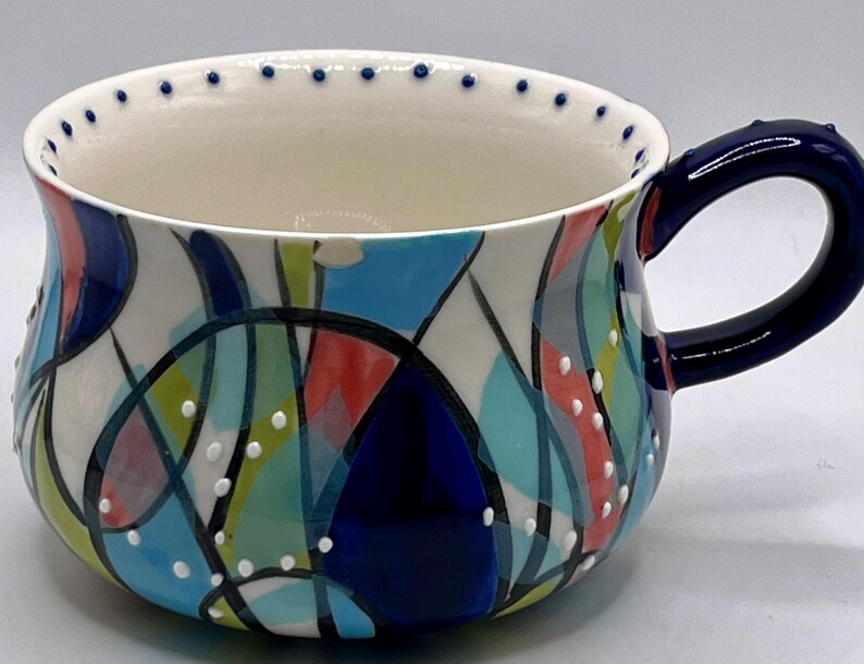 Może przedstawiać: Ceramiczny kubek z ciemnoniebieskim uchwytem i kolorowym abstrakcyjnym wzorem. Kubek ma białe wnętrze i obramowanie ozdobione małymi niebieskimi kropkami. Zewnętrzna strona zdobiona jest mieszanką niebieskich, zielonych, czerwonych i białych kształt&oacute;w.