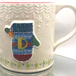 May include: Cream-colored ceramic mug with a red interior and a mitten design. The mitten is decorated with blue, green, and red patterns and the letter "D". The mug has a handle and snowflake and leaf patterns.