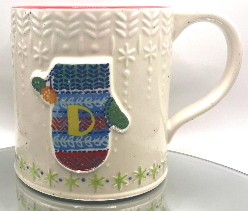May include: Cream-colored ceramic mug with a red interior and a mitten design. The mitten is decorated with blue, green, and red patterns and the letter "D". The mug has a handle and snowflake and leaf patterns.