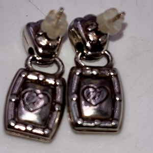 May include: A pair of silver-coloured drop earrings. Each earring has a heart design inside a rectangular frame. The earrings have post-style backs for pierced ears. The metal has a slightly tarnished appearance.