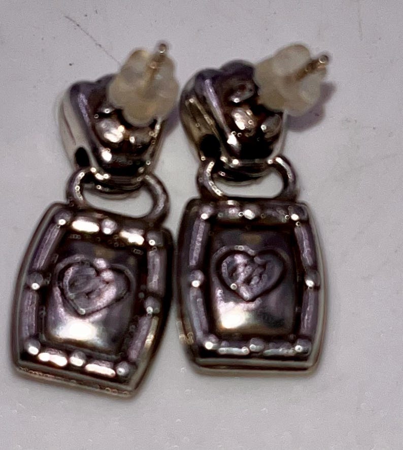 May include: A pair of silver-coloured drop earrings. Each earring has a heart design inside a rectangular frame. The earrings have post-style backs for pierced ears. The metal has a slightly tarnished appearance.