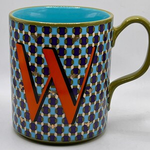 May include: A ceramic mug with a teal interior and a green handle. The exterior features a geometric pattern in blue, purple, and olive green, with an orange letter "W" and gold bee accents. The mug is designed for hot beverages.