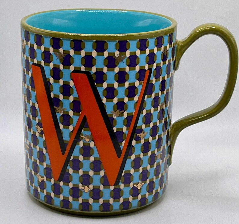 May include: A ceramic mug with a teal interior and a green handle. The exterior features a geometric pattern in blue, purple, and olive green, with an orange letter "W" and gold bee accents. The mug is designed for hot beverages.