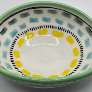 May include: A small, hand-painted ceramic bowl with a light green rim. The interior features a white base with a pattern of blue, green, and yellow shapes, and a black dotted border. This decorative bowl is ideal for serving snacks or as a decorative piece.