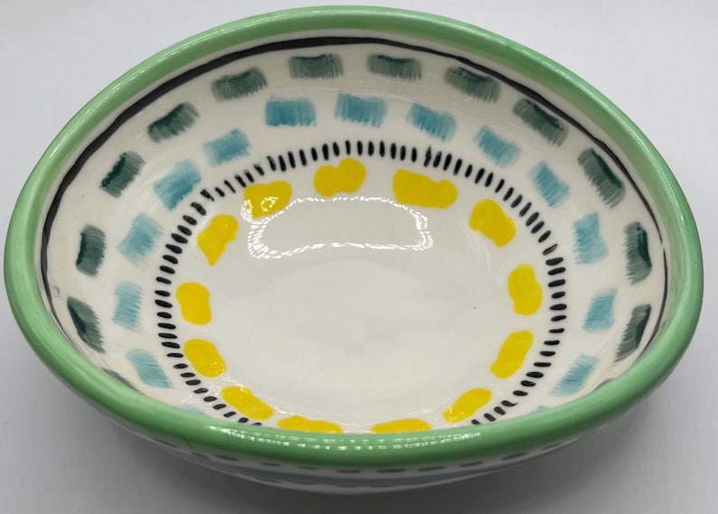 May include: A small, hand-painted ceramic bowl with a light green rim. The interior features a white base with a pattern of blue, green, and yellow shapes, and a black dotted border. This decorative bowl is ideal for serving snacks or as a decorative piece.