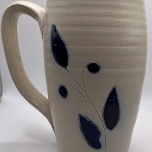 May include: A handmade ceramic mug with a cream-colored base and a large handle. The mug features a dark blue leaf and berry design. The mug is approximately 6 inches tall.