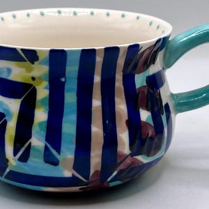 May include: A ceramic mug with a teal handle and a white interior. The exterior features a colorful design of blue stripes, teal and yellow shapes, and maroon leaf-like patterns. The rim has small teal dots.