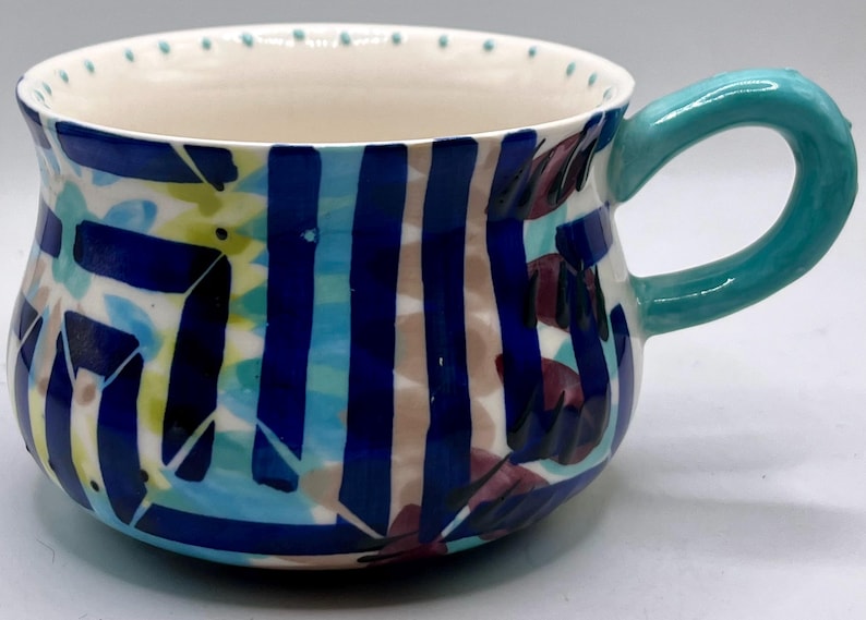 May include: A ceramic mug with a teal handle and a white interior. The exterior features a colorful design of blue stripes, teal and yellow shapes, and maroon leaf-like patterns. The rim has small teal dots.
