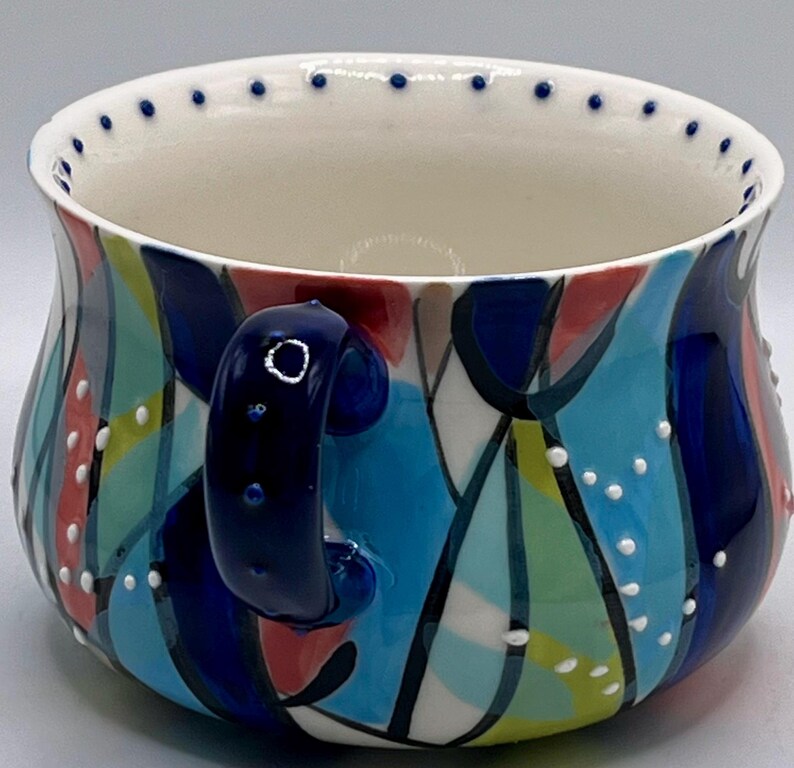 Może przedstawiać: Ceramiczny kubek z kolorowym abstrakcyjnym wzorem. Kubek ma białe wnętrze i ciemnoniebieski uchwyt. Zewnętrzna strona zdobiona jest niebieskimi, czerwonymi, zielonymi i ż&oacute;łtymi kształtami, zaakcentowanymi białymi kropkami. Obrzeże wykończone jest małymi niebieskimi kropkami.