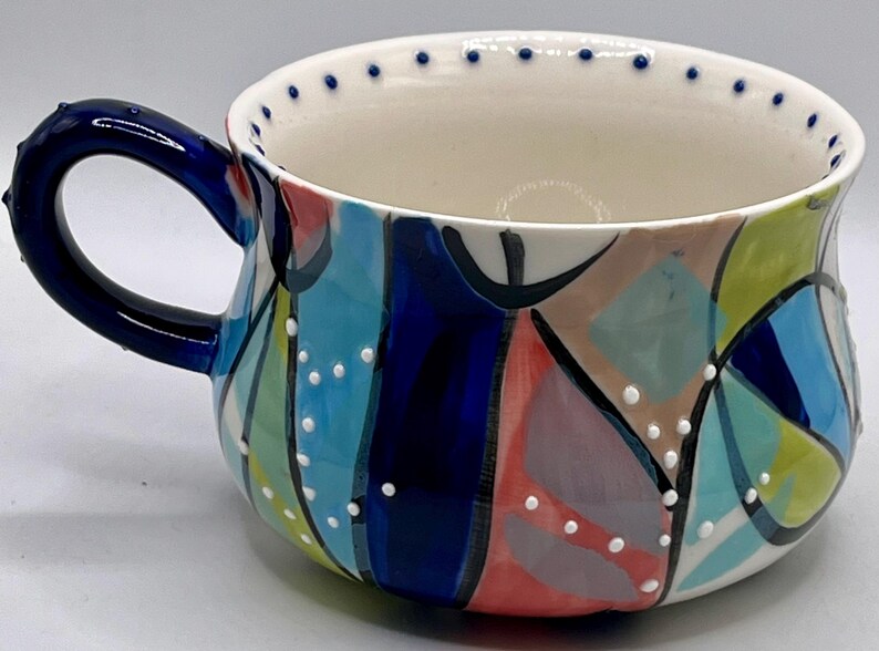 Może przedstawiać: Ceramiczny kubek z ciemnoniebieskim uchwytem i kolorowym abstrakcyjnym wzorem. Kubek ma białe wnętrze i obrzeże ozdobione małymi niebieskimi kropkami. Zewnętrzna strona pomalowana jest na niebiesko, turkusowo, r&oacute;żowo i zielono, z białymi kropkami.
