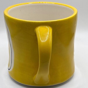 Puede incluir: Una taza de cer&aacute;mica amarilla con un asa curva. La taza tiene un interior blanco y un dise&ntilde;o en el lateral con detalles en blanco y negro. La taza est&aacute; sobre una superficie blanca.