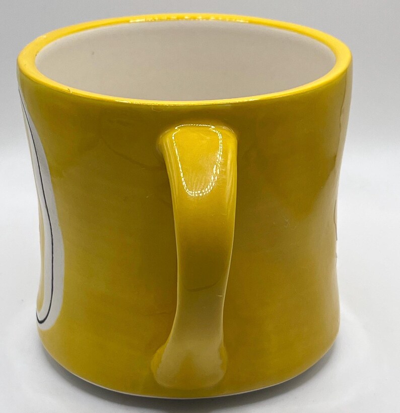 Puede incluir: Una taza de cer&aacute;mica amarilla con un asa curva. La taza tiene un interior blanco y un dise&ntilde;o en el lateral con detalles en blanco y negro. La taza est&aacute; sobre una superficie blanca.