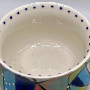 Może przedstawiać: Ceramiczna miska z kremowym wnętrzem i kolorowym, geometrycznym wzorem na zewnątrz. Obręcz jest ozdobiona małymi niebieskimi kropkami. Projekt miski zawiera odcienie niebieskiego, zielonego i czerwonego.