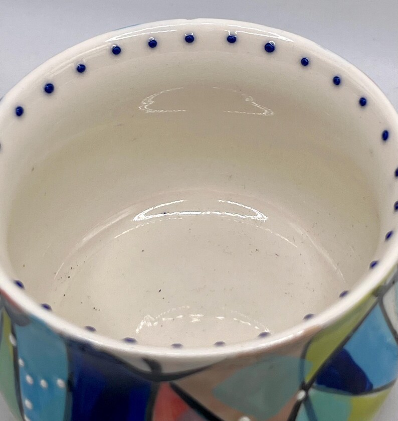 Może przedstawiać: Ceramiczna miska z kremowym wnętrzem i kolorowym, geometrycznym wzorem na zewnątrz. Obręcz jest ozdobiona małymi niebieskimi kropkami. Projekt miski zawiera odcienie niebieskiego, zielonego i czerwonego.