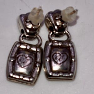 May include: A pair of silver-coloured earrings. Each earring has a rectangular pendant with a heart design in the centre. The earrings have a post-back closure. The earrings are likely made of metal and are designed for pierced ears.