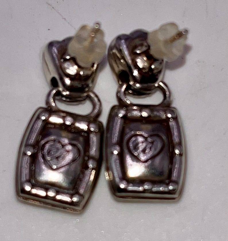 May include: A pair of silver-coloured earrings. Each earring has a rectangular pendant with a heart design in the centre. The earrings have a post-back closure. The earrings are likely made of metal and are designed for pierced ears.