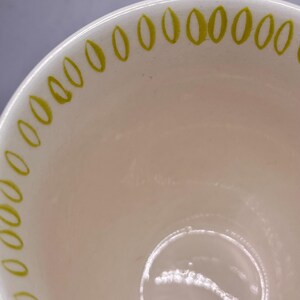 May include: Cream-colored ceramic bowl with a glossy finish. The rim is decorated with a repeating pattern of small, olive-green ovals. The interior of the bowl is a soft, off-white color, and the bowl appears to be empty.