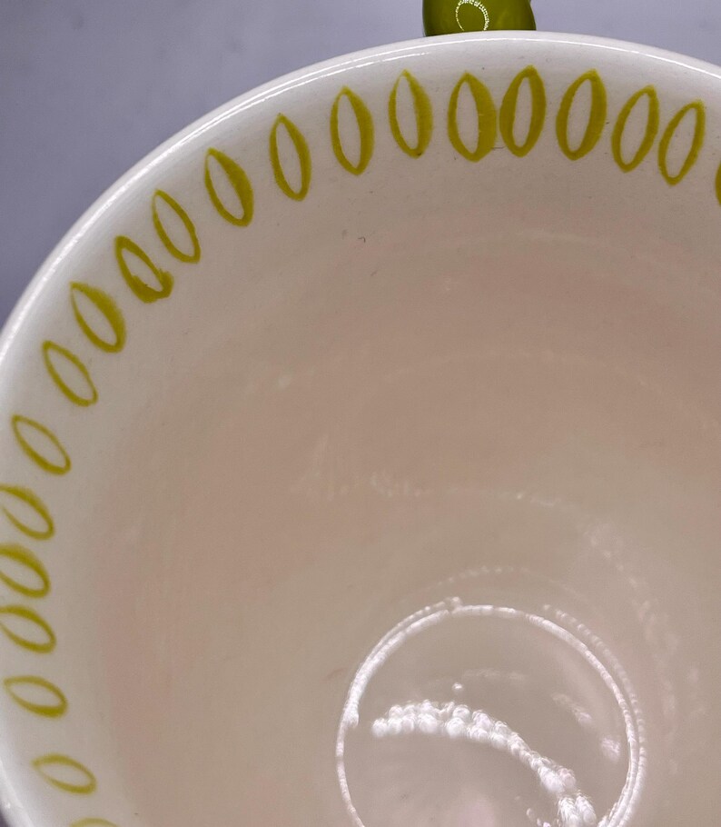 May include: Cream-colored ceramic bowl with a glossy finish. The rim is decorated with a repeating pattern of small, olive-green ovals. The interior of the bowl is a soft, off-white color, and the bowl appears to be empty.